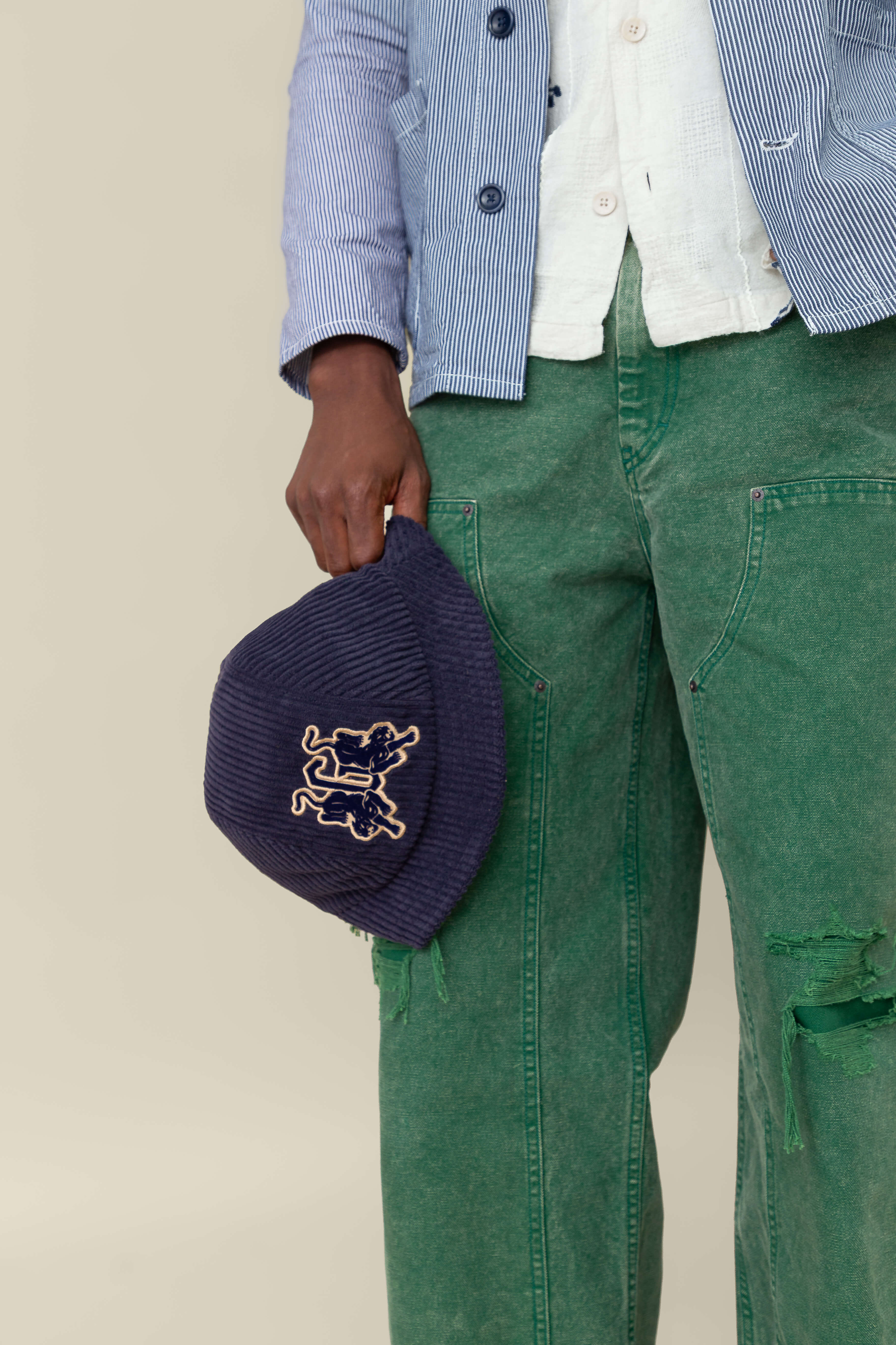 A person wearing green jeans and a blue striped jacket holds the "Yes Coach" corduroy bucket hat by Goorin Bros.
