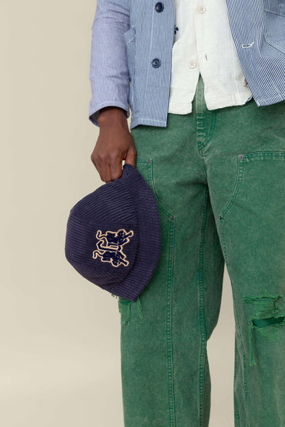 A person wearing green jeans and a blue striped jacket holds the "Yes Coach" corduroy bucket hat by Goorin Bros.