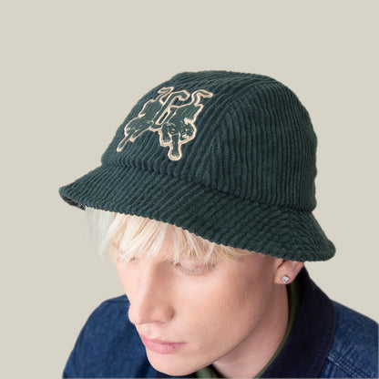 A person with platinum blonde hair wears the "Yes Coach" textured corduroy bucket hat by Goorin Bros., featuring two embroidered dog illustrations, paired with a denim jacket against a plain light background.