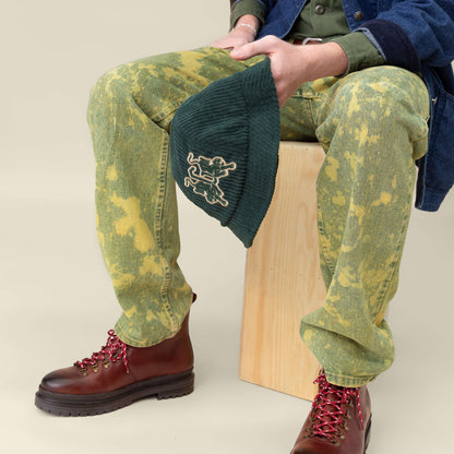 Seated on a wooden block, this person holds the Yes Coach bucket hat by Goorin Bros., and wears green acid-wash pants with brown boots featuring red laces.