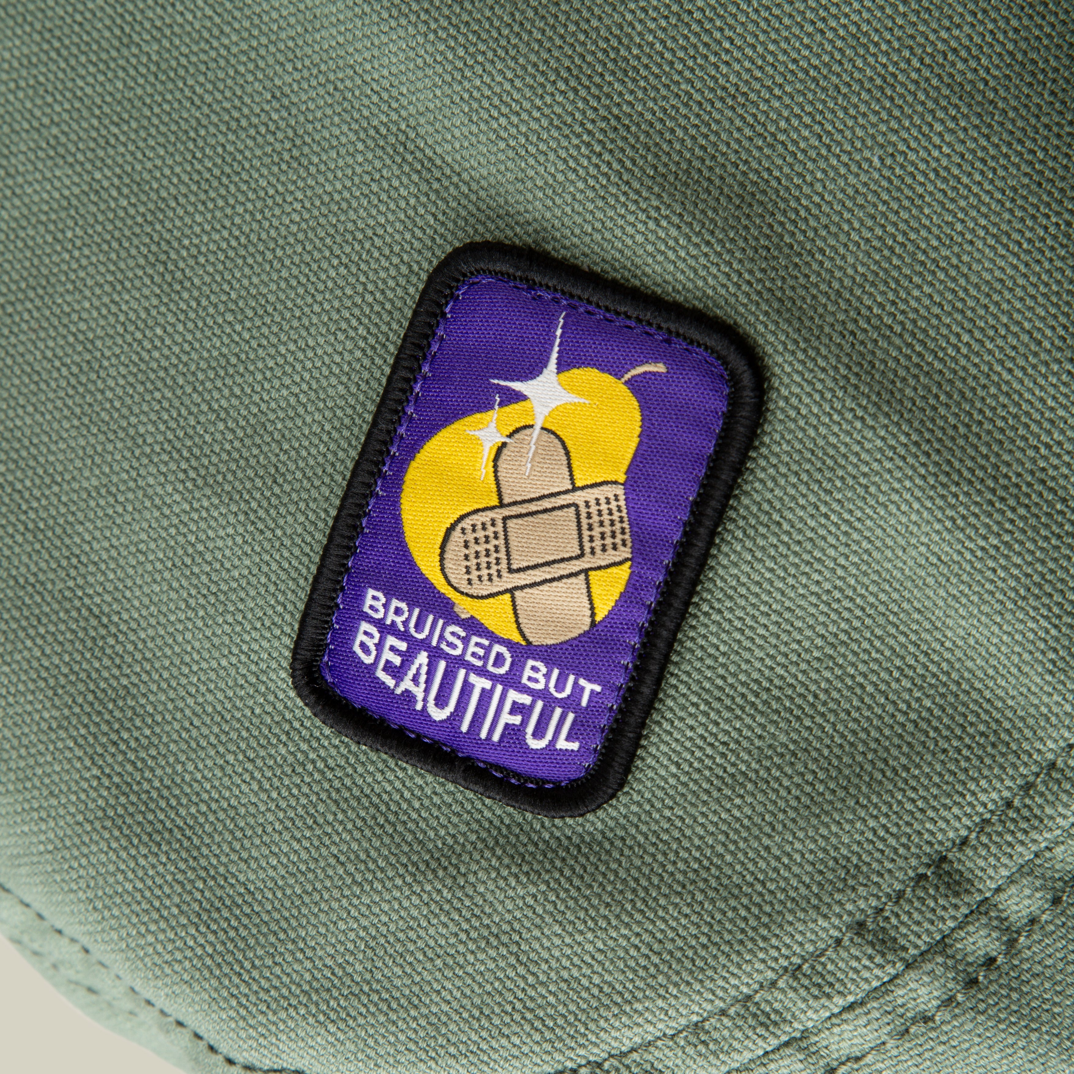 The Goorin Bros. PLU 1895 patch on olive fabric shows two bandages over a bruised yellow fruit with "BRUISED BUT BEAUTIFUL" on purple—a unique souvenir or fun addition to your bulletin board.