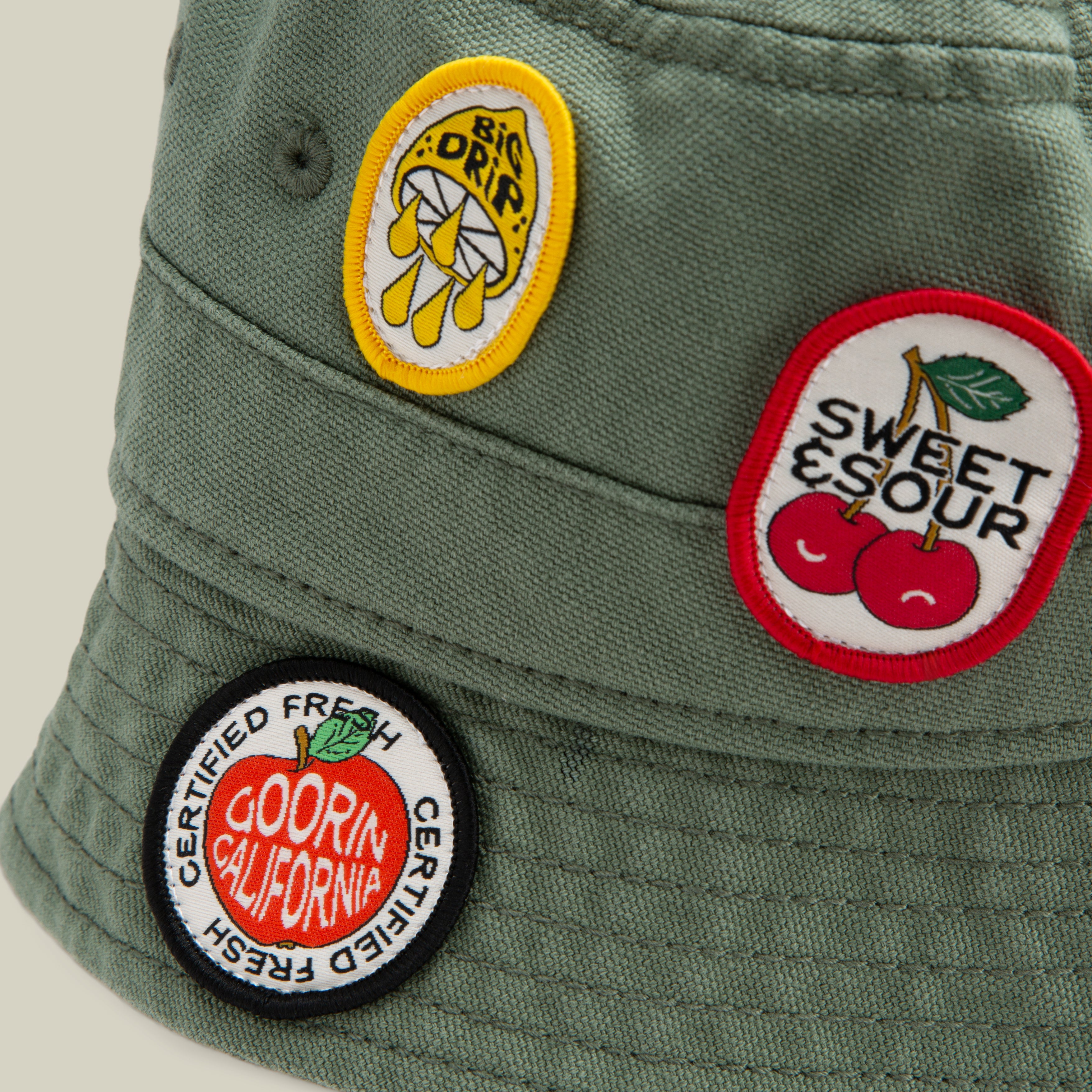 The Goorin Bros. PLU 1895 is an olive green bucket hat souvenir with three colorful embroidered patches: a lemon with BIG DRIP, cherries with SWEET & SOUR, and a round DOORIN CALIFORNIA CERTIFIED FRESH patch.