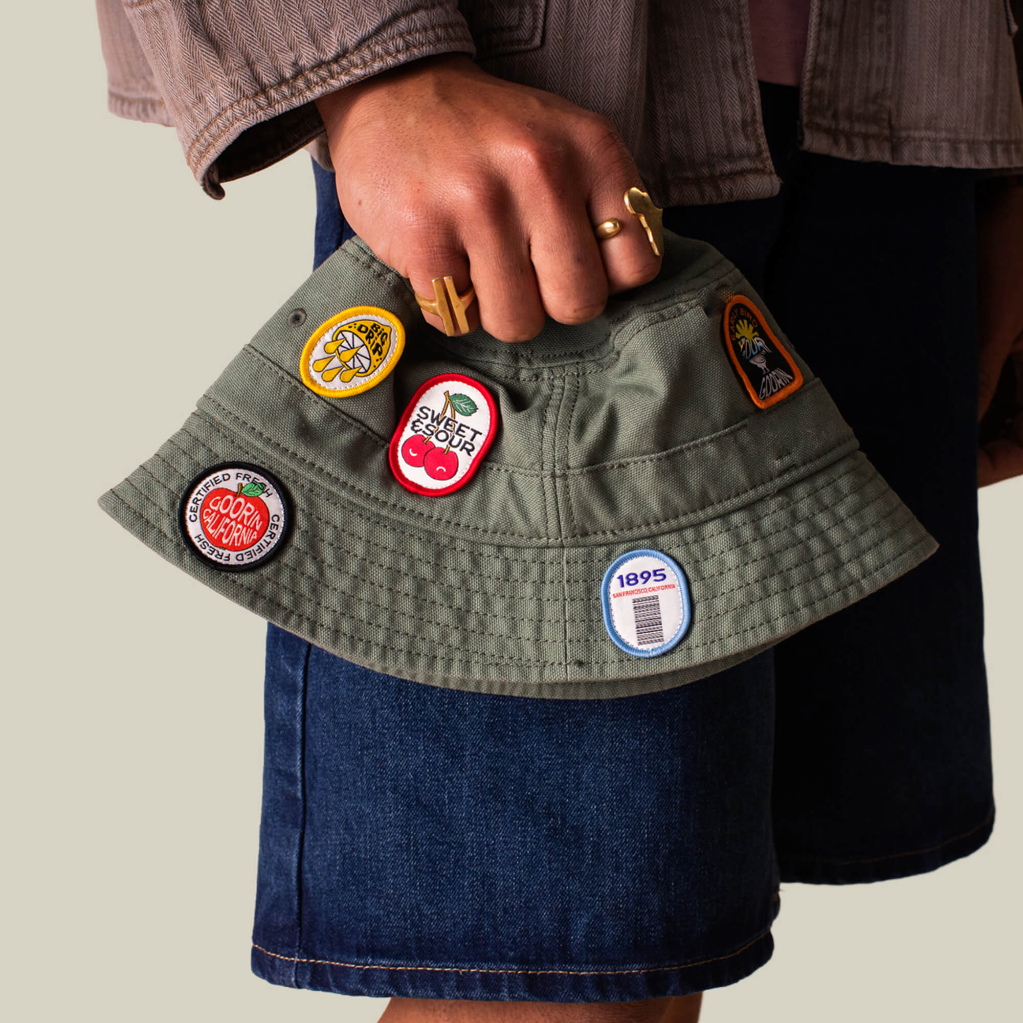 A person in denim shorts holds the olive green PLU 1895 bucket hat from Goorin Bros., featuring colorful embroidered patches.