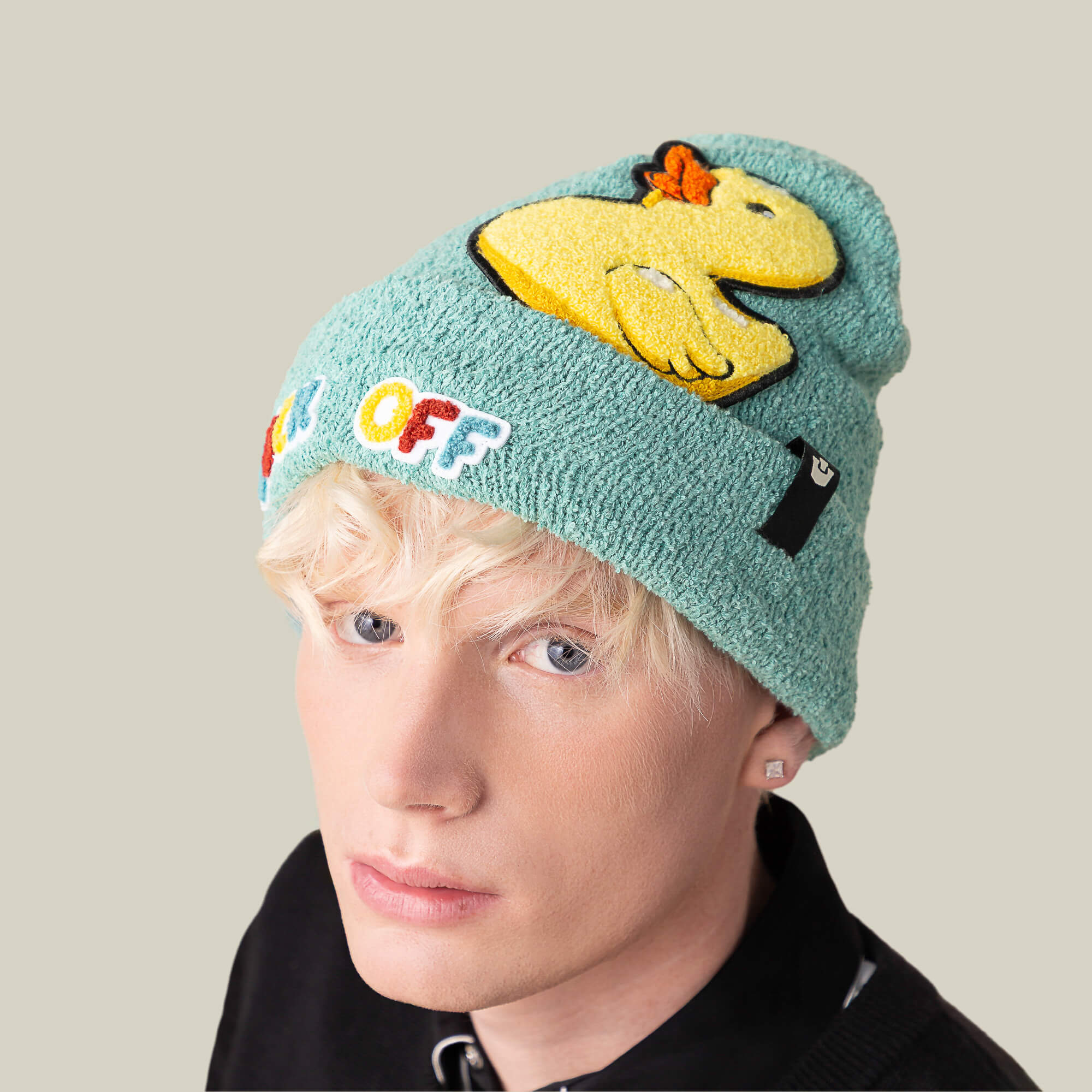 A person with light blond hair wears the "Awfully Fond of You" turquoise warm beanie by Goorin Bros., featuring a yellow duck patch and colorful OFF lettering, set against a plain beige background.