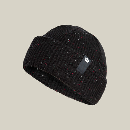 The No Comply beanie by Goorin Bros. is a black ribbed knit hat with multicolored flecks and a small black rectangular patch on the folded brim, shown against a plain beige background.