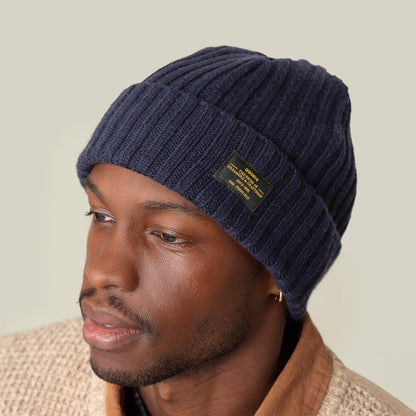A man wears the navy blue Dream Cap by Goorin Bros.—a luxurious cashmere wool knitted beanie with a black label—paired with a beige textured jacket and brown shirt against a neutral background.