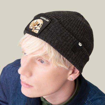A person with light blond hair wears the Goorin Bros. Cancelled Beanie—a charcoal knit hat with a skull patch and "CANCELLED" on the cuff—styled with a denim jacket and stud earrings.
