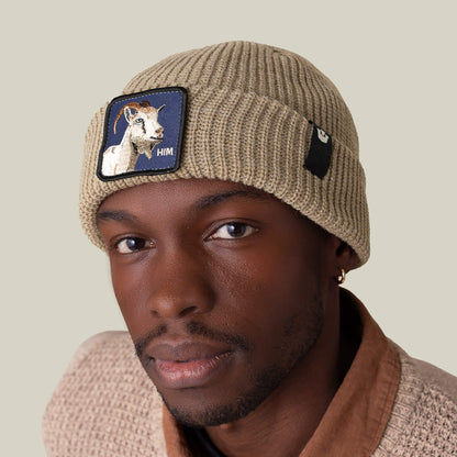 A person wears The Him Beanie by Goorin Bros.—a cozy tan knit hat with a goat patch—paired with a textured beige sweater, looking at the camera against a plain background.