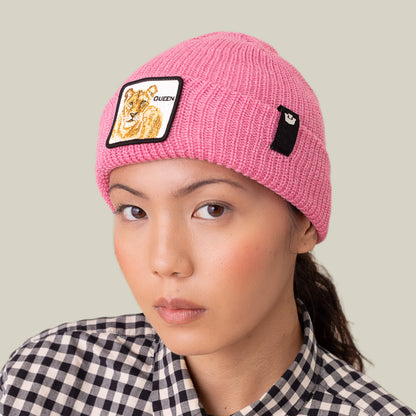A person wears Goorin Bros.' The Queen Beanie, a pink knit winter hat with a lioness patch and QUEEN text, paired with a black-and-white checkered shirt against a plain background.