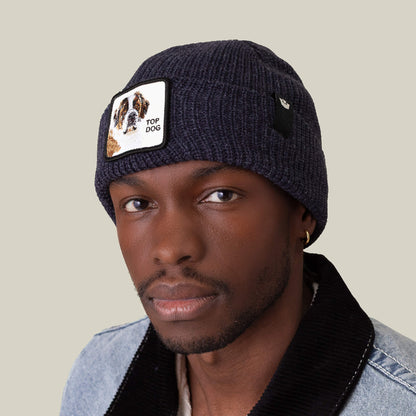 A man wears The Top Beanie by Goorin Bros., a navy knit hat featuring a St. Bernard dog patch, paired with a denim jacket with a black collar.