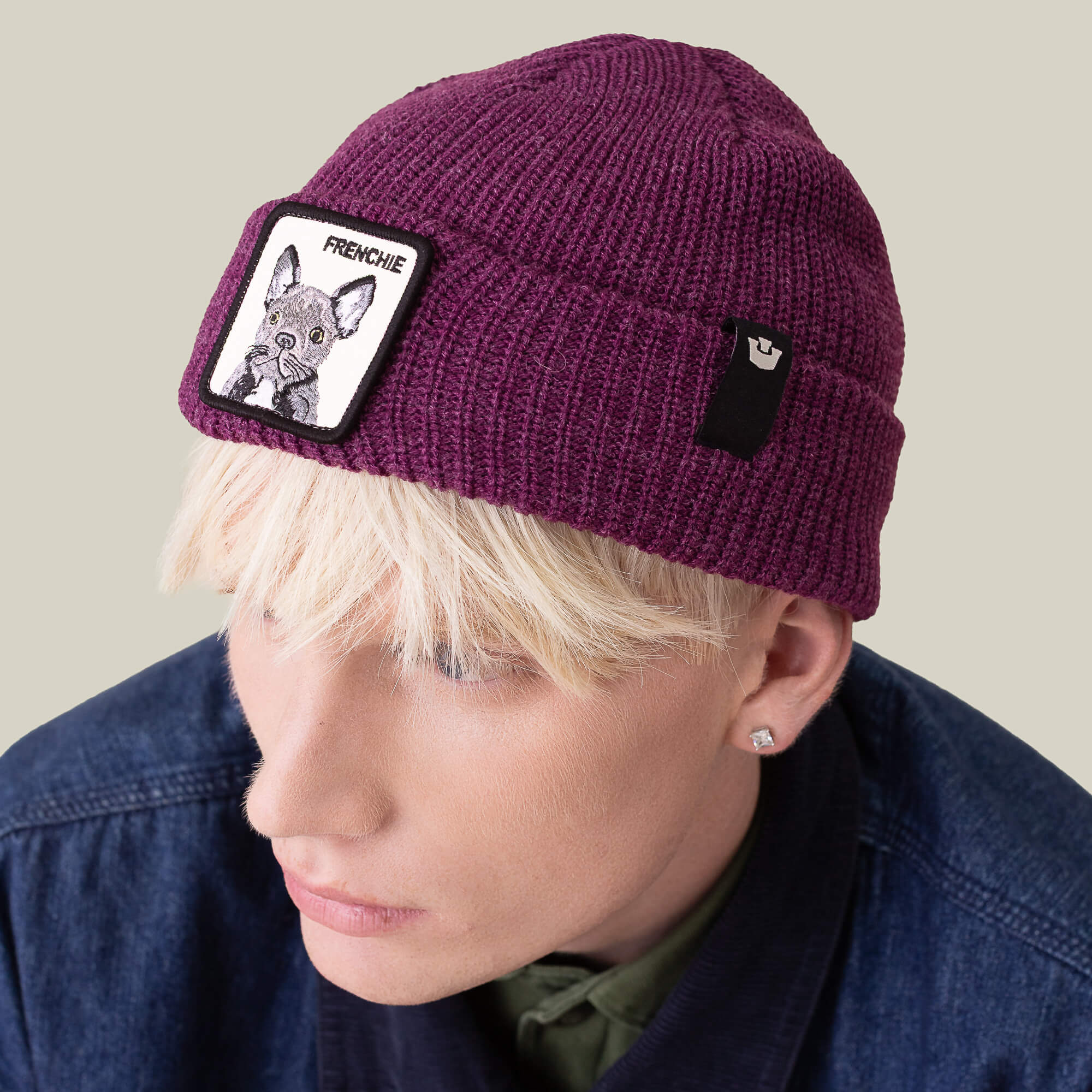 A person with short blonde hair wears The Frenchie Beanie by Goorin Bros.—a plum knit hat with a French Bulldog patch—paired with a dark denim jacket for cozy street style.
