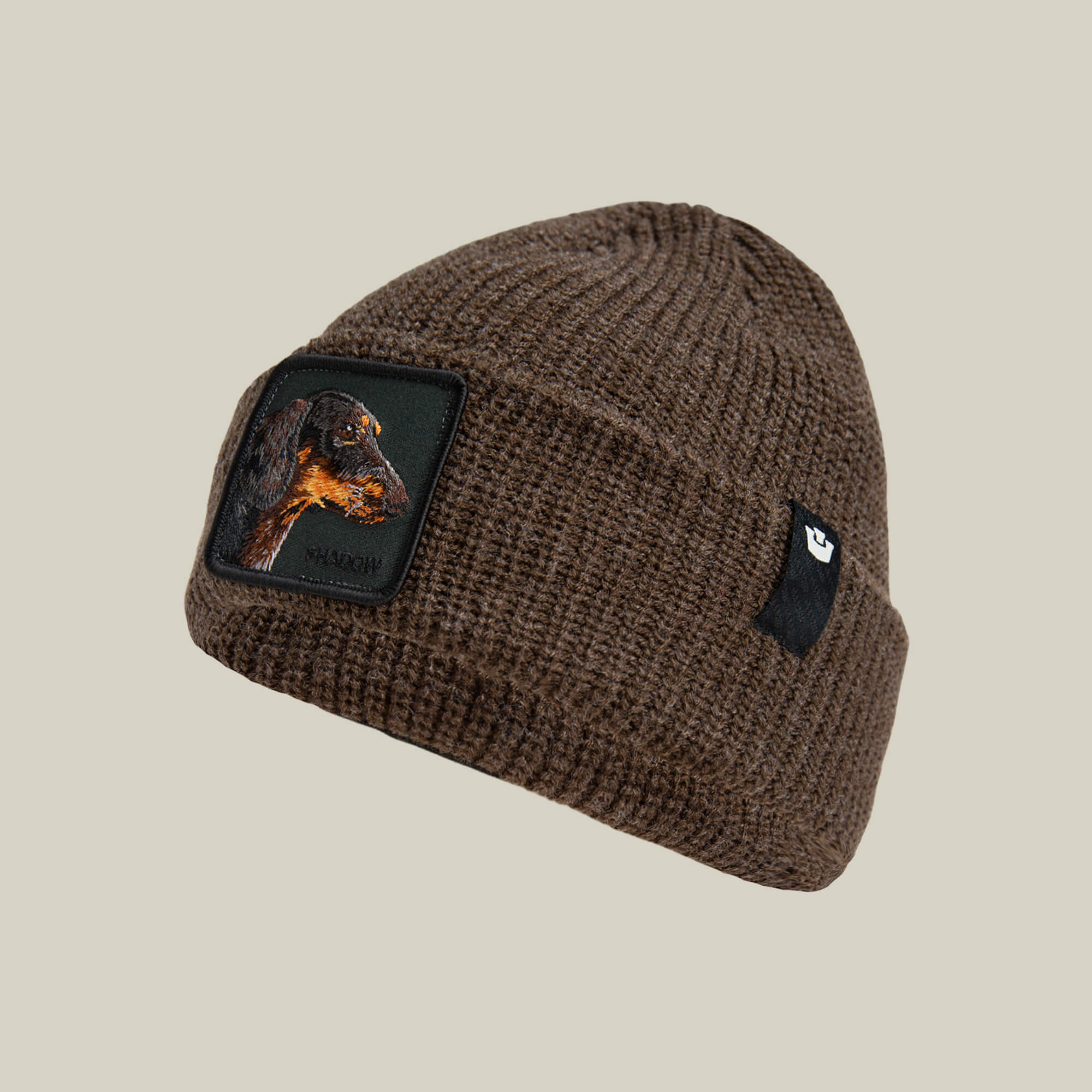 The Shadow Beanie by Goorin Bros. is a dark brown knit beanie featuring a dachshund patch on the side and a small black logo tag, shown on a plain beige background.