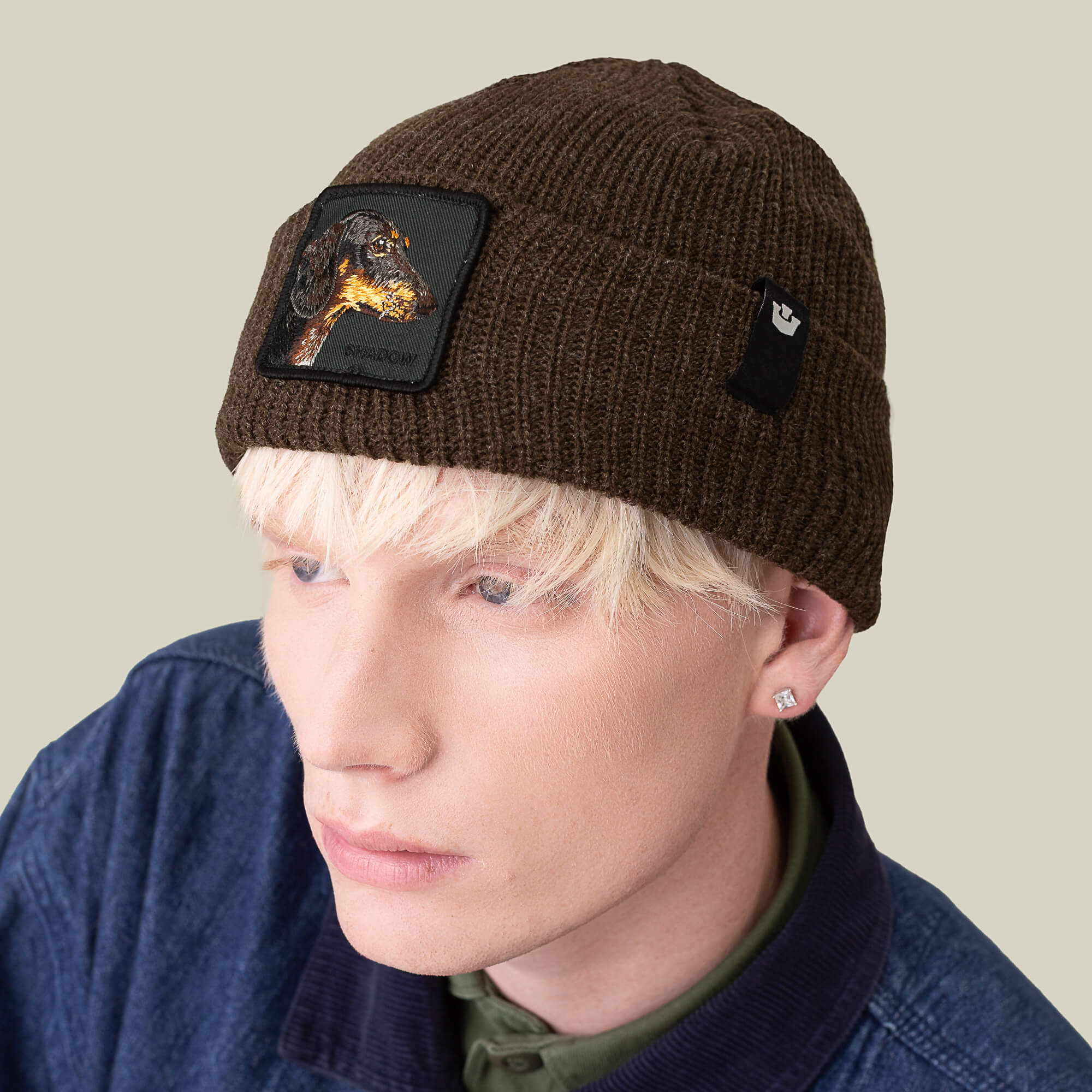 A person with light blond hair wears the Goorin Bros. Shadow Beanie—a dark brown knit hat with a dachshund patch—paired with a blue jacket over a green shirt, set against a plain light background.
