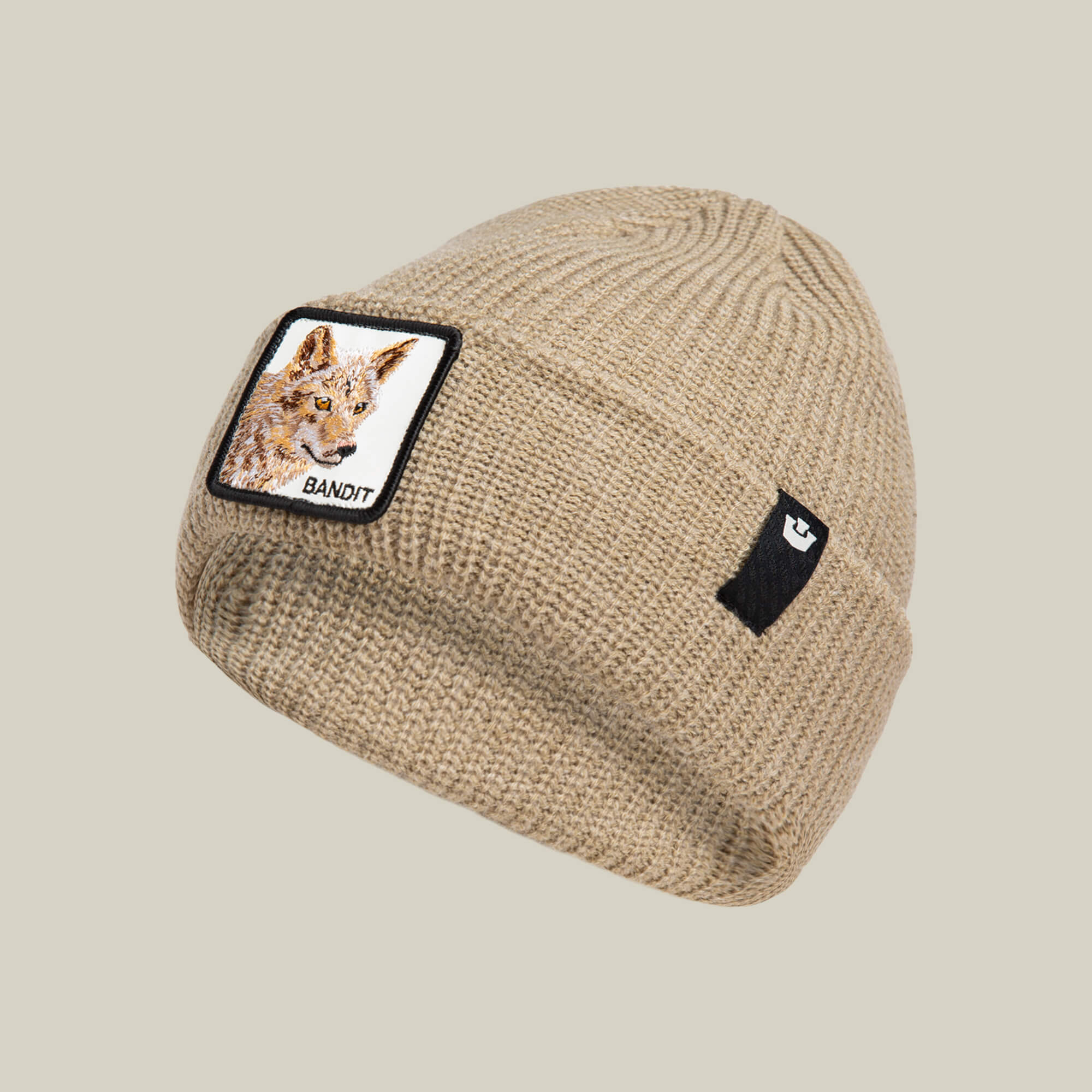 The Bandit Beanie by Goorin Bros. is an earth-tone knit winter hat featuring a coyote illustration patch labeled "Bandit" and a small black logo tag on the cuff, shown against a plain background.