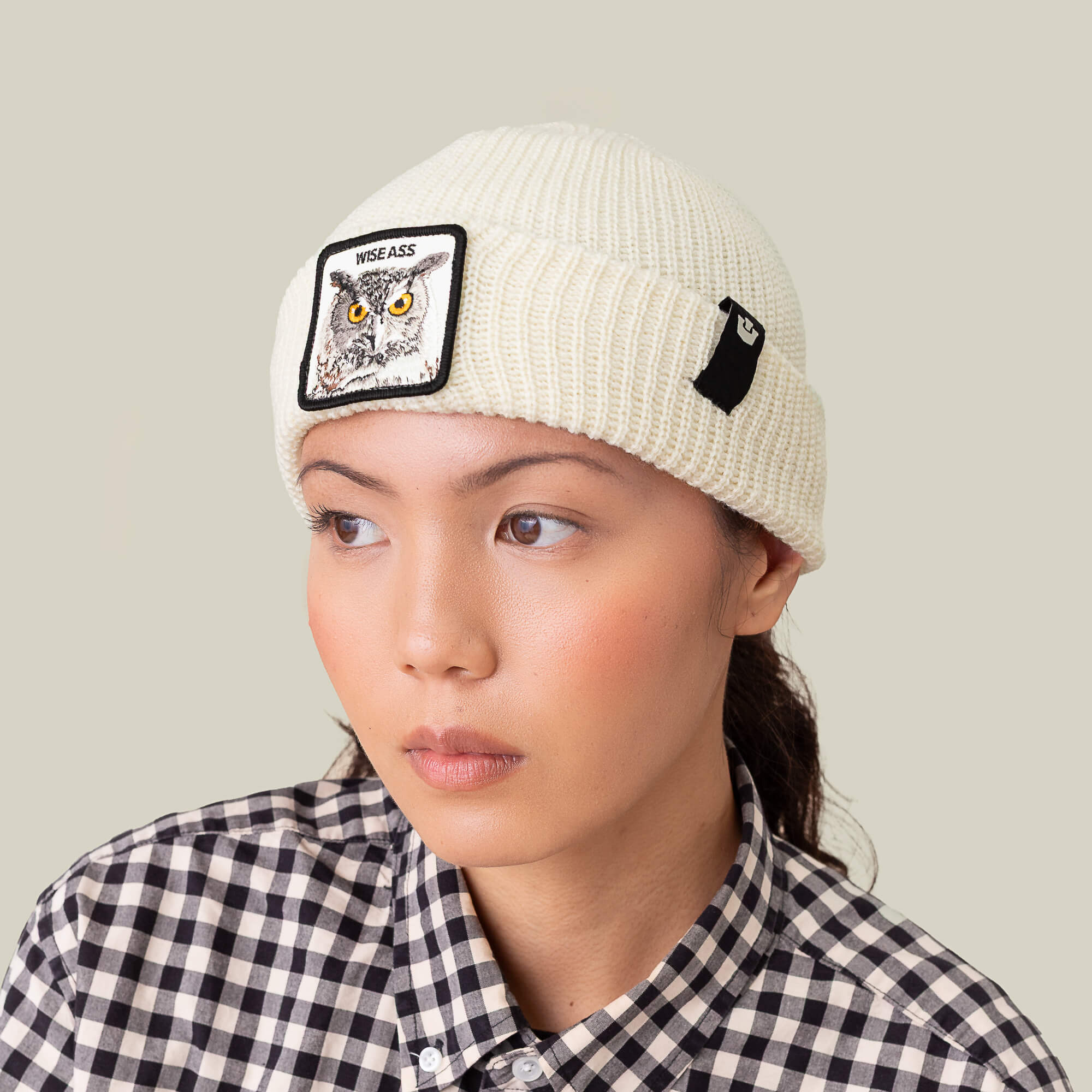 A person in the Goorin Bros. Wise Ass Beanie, featuring an owl patch, styled with a black-and-white checkered shirt and looking slightly right—showcasing a fun winter accessory statement.