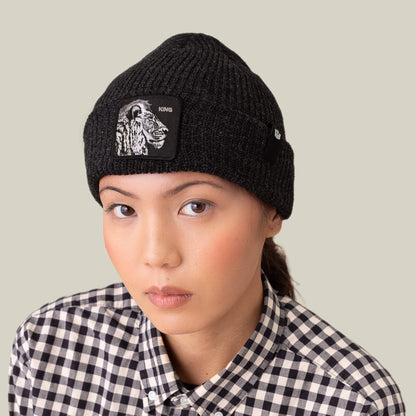 A person wears The Lion Beanie by Goorin Bros.—a black cuffed beanie with a patch—paired with a black-and-white checkered shirt, looking directly at the camera against a plain background.