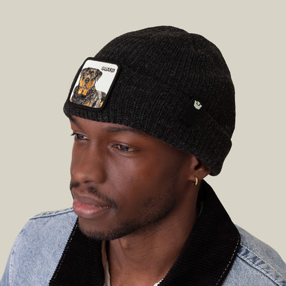 A man wears The Guard Beanie by Goorin Bros., a black knit hat featuring a Rottweiler patch and "GUARD," styled with a light denim jacket with a black collar.