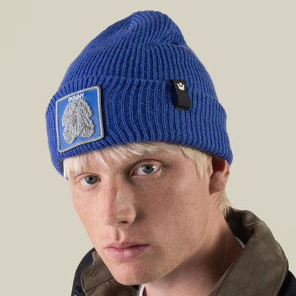 A person wearing a blue rib-knit cuffed Double Stuffed - Bunny Beanie featuring a patch of a gray rabbit and the word BUNNY.