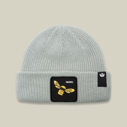 The Queen Bee Beanie by Goorin Bros is a light gray ribbed knit beanie featuring a black square patch with gold embroidered bee and "QUEEN" text on the front, displayed against a neutral background.