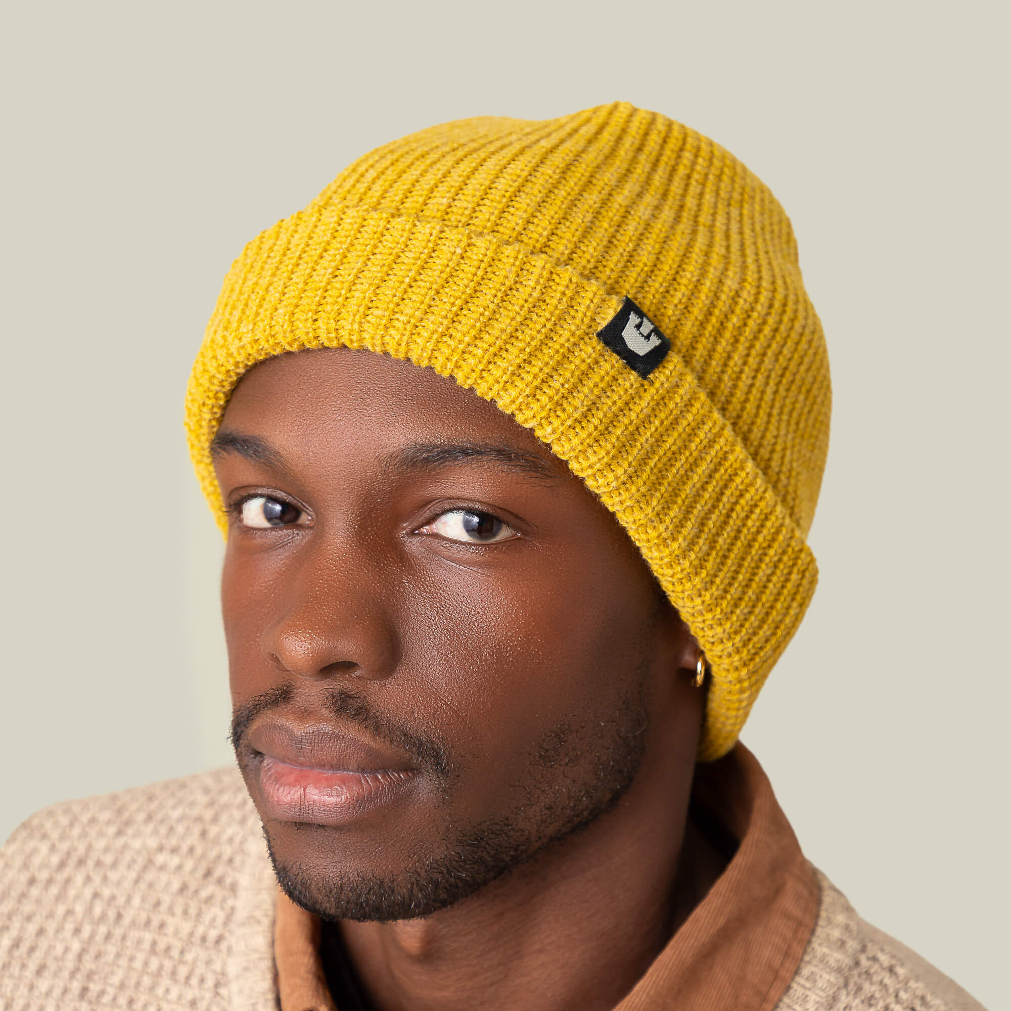 A person wears a yellow Goorin Bros. Air to Fakie beanie made of lightweight poly knit with a tan textured jacket, looking directly at the camera against a plain light background—an all-season style.