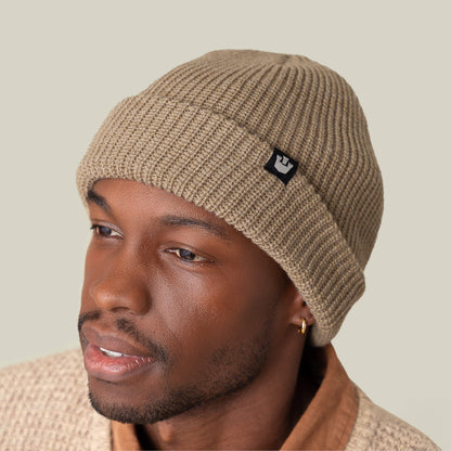 Someone wears the Goorin Bros. Air to Fakie beige Watchman beanie with a black logo and a matching textured sweater, glancing sideways against a plain background. Its lightweight poly knit makes it ideal for all-season use.