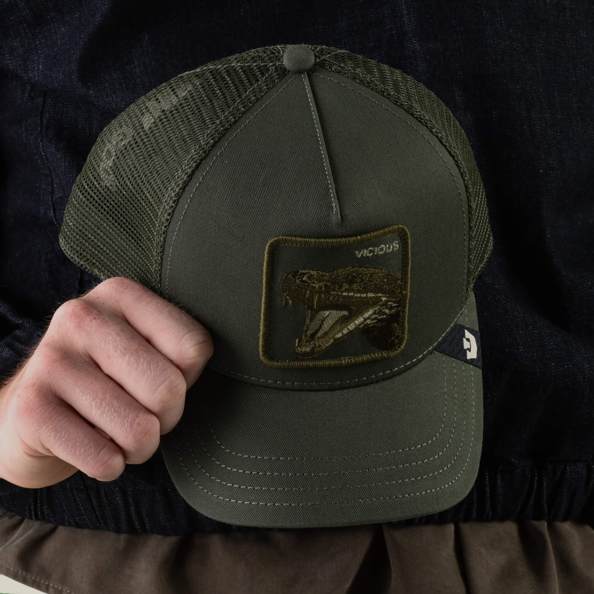 A person holding the Goorin Bros. Circle of Life - Vicious trucker hat, featuring an olive front, green mesh back, and a patch with an open-mouthed snake and the word “VICIOUS”. This stylish cap also includes an adjustable snapback for a comfortable fit.