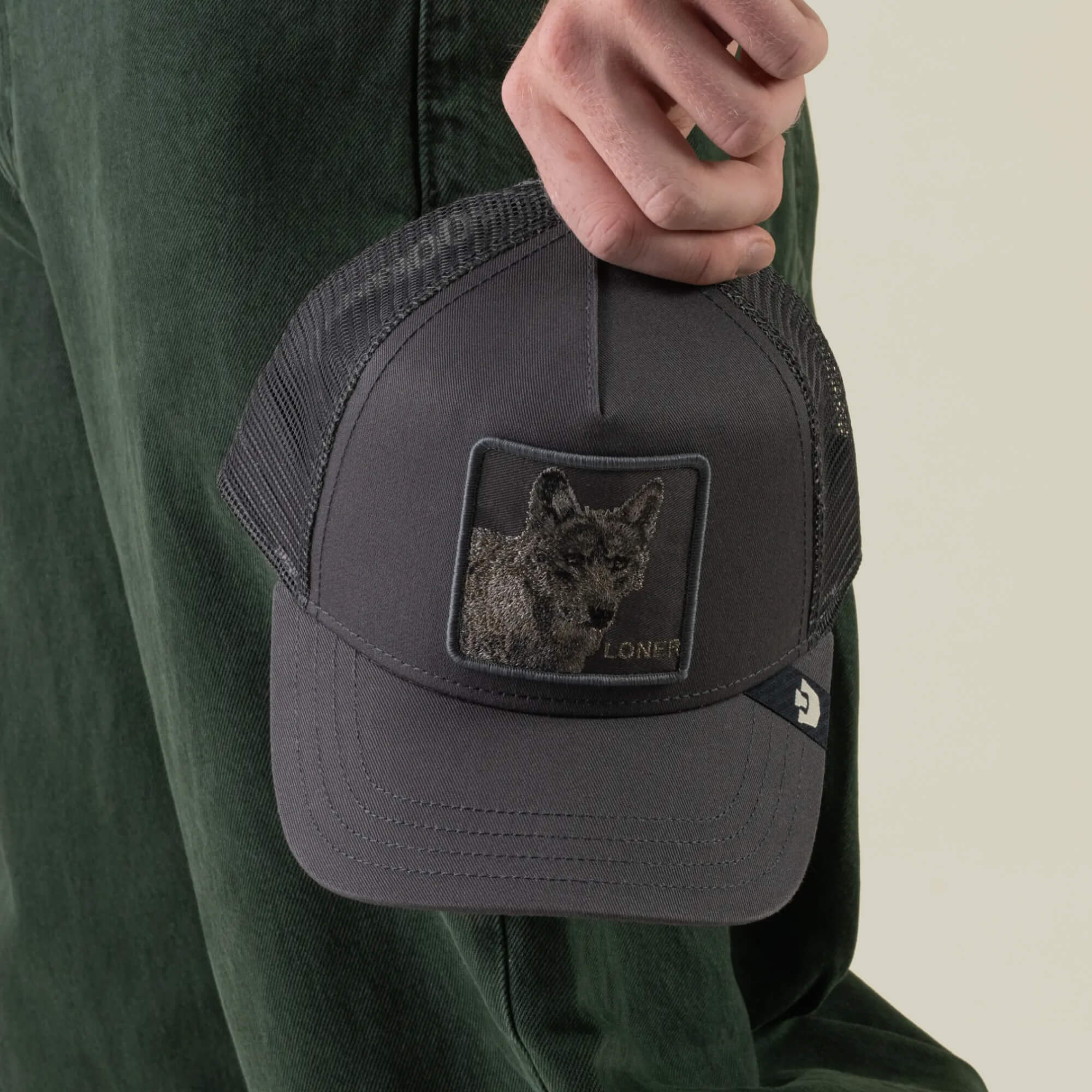 A person holding the Goorin Bros. “Circle of Life - Loner” dark gray embroidered wolf snapback trucker hat, standing next to green pants.