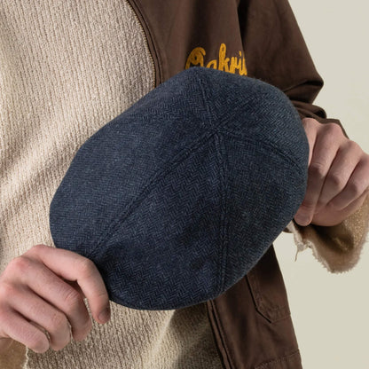 A person holds the Goorin Bros. “Harry Bones” dark blue flat cap in both hands, while wearing a beige sweater and a brown jacket with partial yellow lettering visible. This heavy-duty headwear is expertly crafted from 100% wool, merging durability with timeless style.