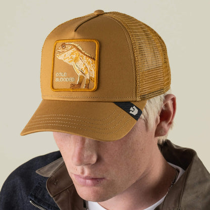 A person wearing the Goorin Bros. Circle of Life - Cold Blooded brown mesh-back trucker hat, featuring a patch with a bearded lizard and the words COLD BLOODED, dressed in a dark jacket over a collared shirt.