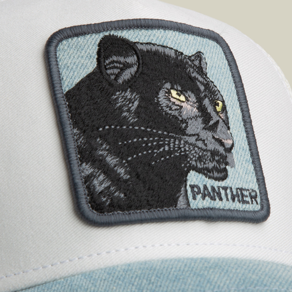 Introducing the Goorin Bros. Denim Panther: a low-profile trucker hat in white and light blue, featuring a denim brim and an embroidered patch with a black panther and “PANTHER” in bold capital letters.