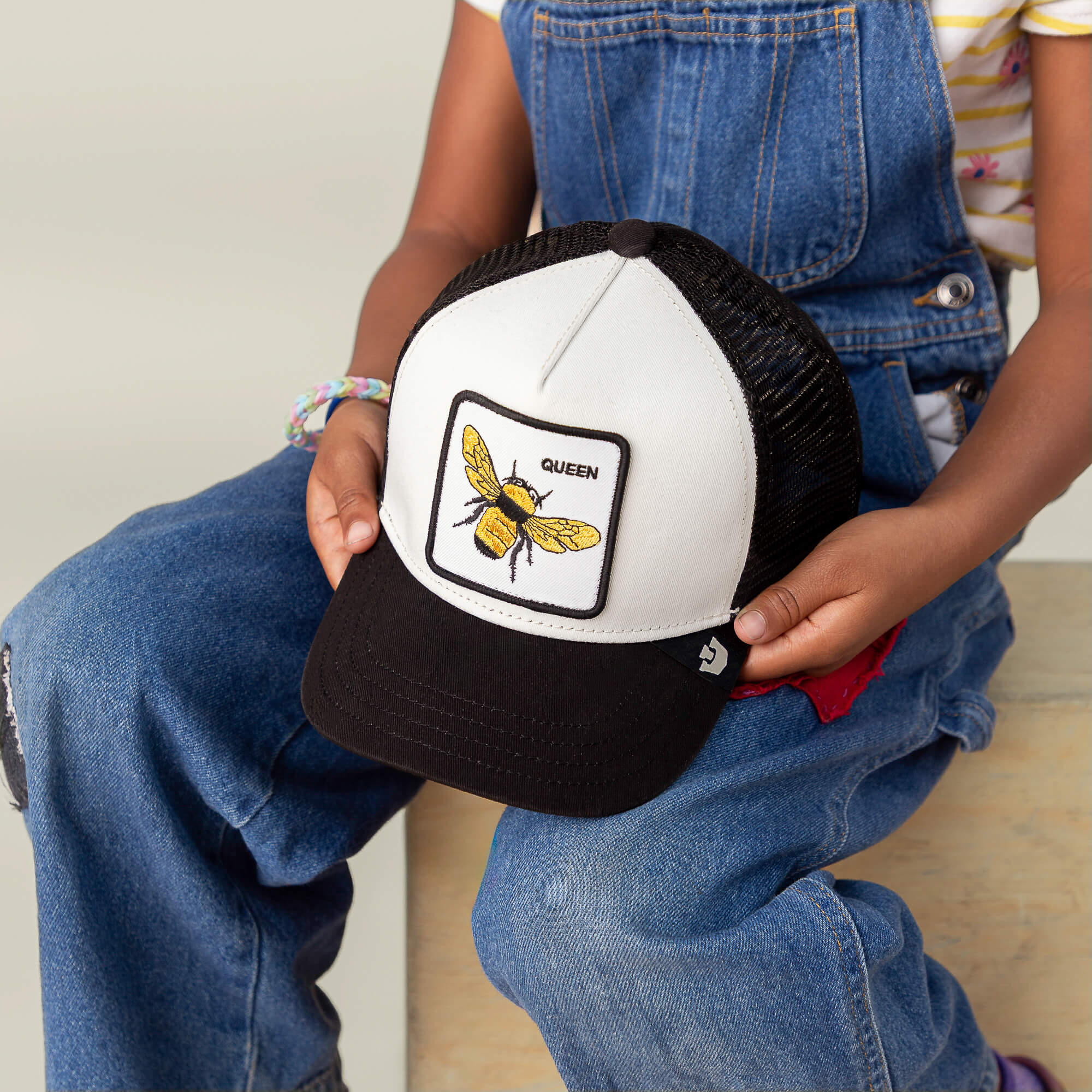 A child in denim overalls holds Goorin Bros.’ The Queen Bee Mini, a black-and-white kids cap featuring an embroidered crowned bee and the word QUEEN.