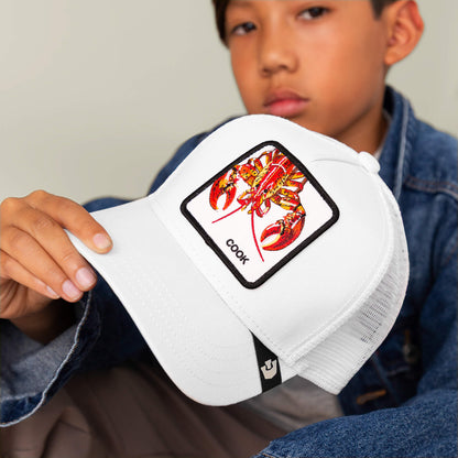 A person in a denim jacket holds the Goorin Bros. Lemme Cook Mini hat, a kid-sized white cap with a patch featuring an illustrated fiery lobster and the word “COOK.”.