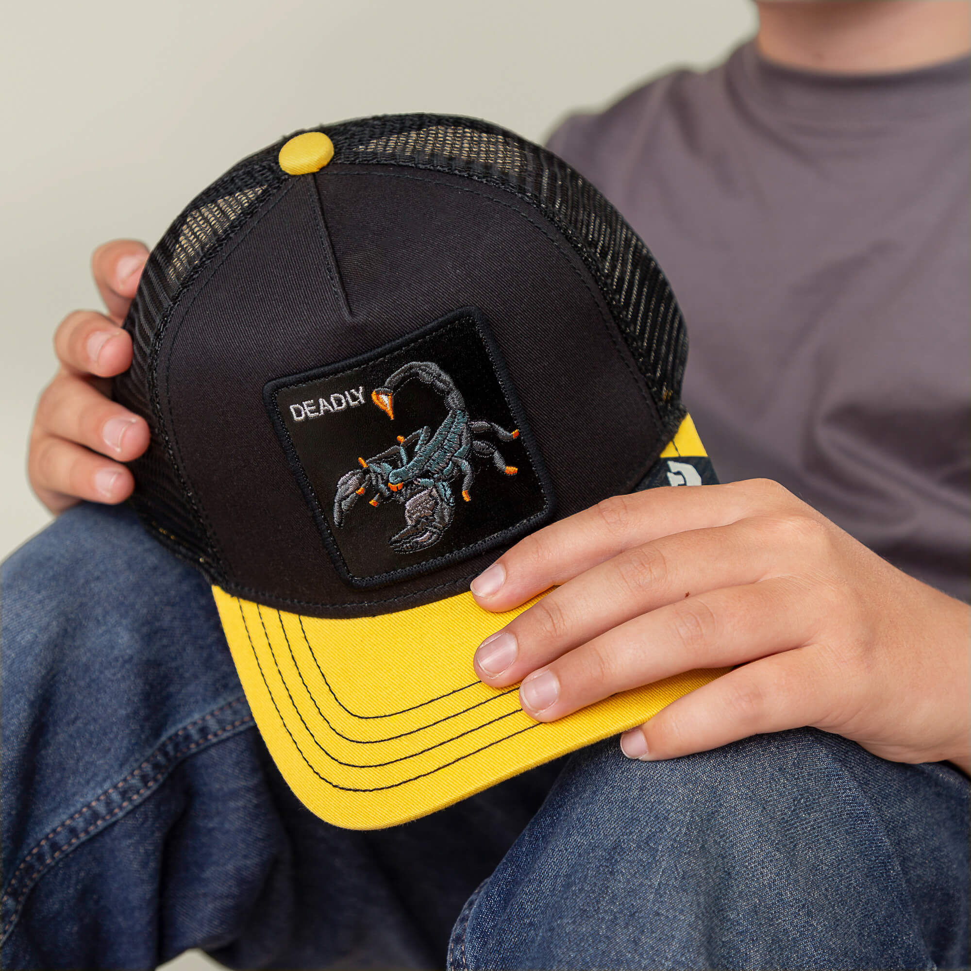 A person in jeans and a gray shirt holds the Goorin Bros. Deadly Mini kids hat, featuring a yellow brim and an embroidered scorpion patch.