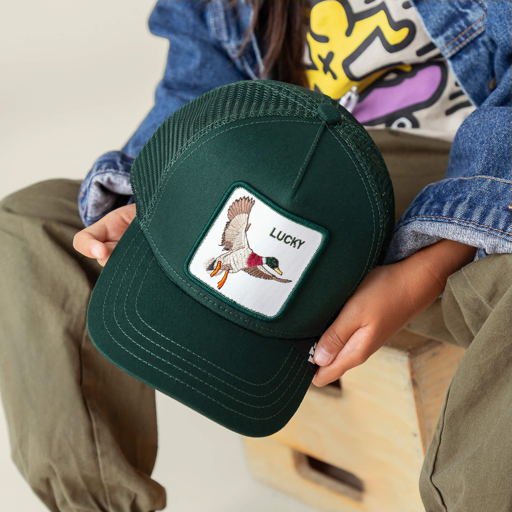 A person holds The Lucky Duck Mini by Goorin Bros., a kid-sized dark green cap with a flying duck patch and "LUCKY" on the crown.