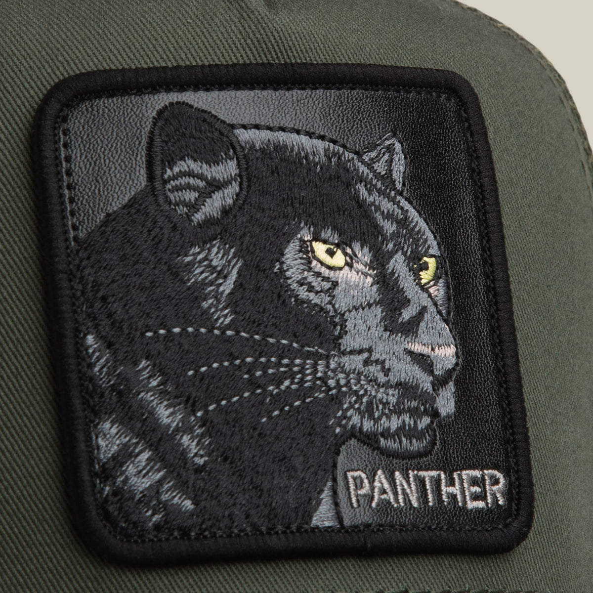 The Goorin Bros. Panther Trucker Mini features an olive green base with a detailed black embroidered panther patch showing yellow eyes, displayed in close-up on the hat's front panel.