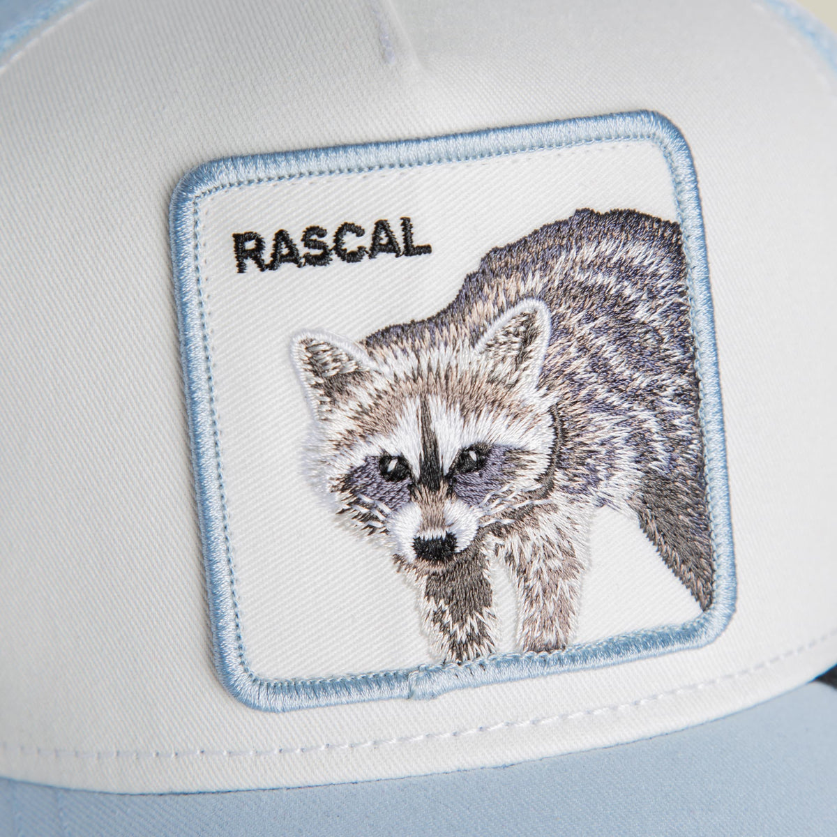 The Goorin Bros. Rascal Raccoon Mini is a white trucker hat with light blue trim featuring an embroidered raccoon patch on the front and the text "RASCAL" above it.