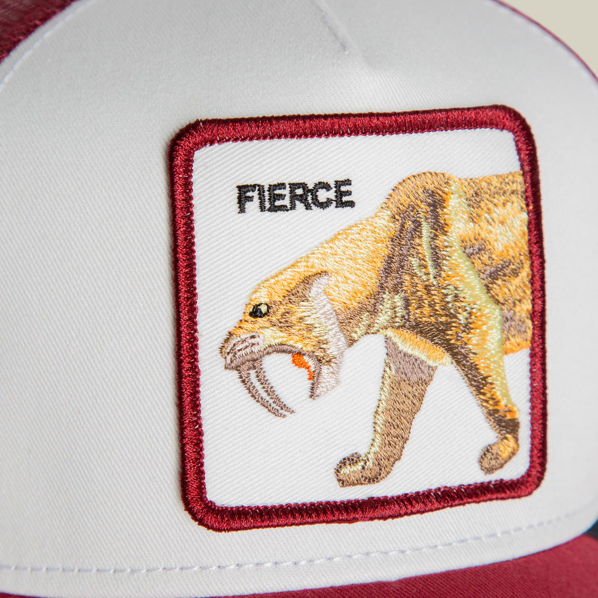 The Goorin Bros. Fierce Saber-tooth Tiger Mini trucker hat features a red and white color block design with an embroidered patch of a saber-tooth tiger with bared fangs on the front panel, shown flat lay on a white background.