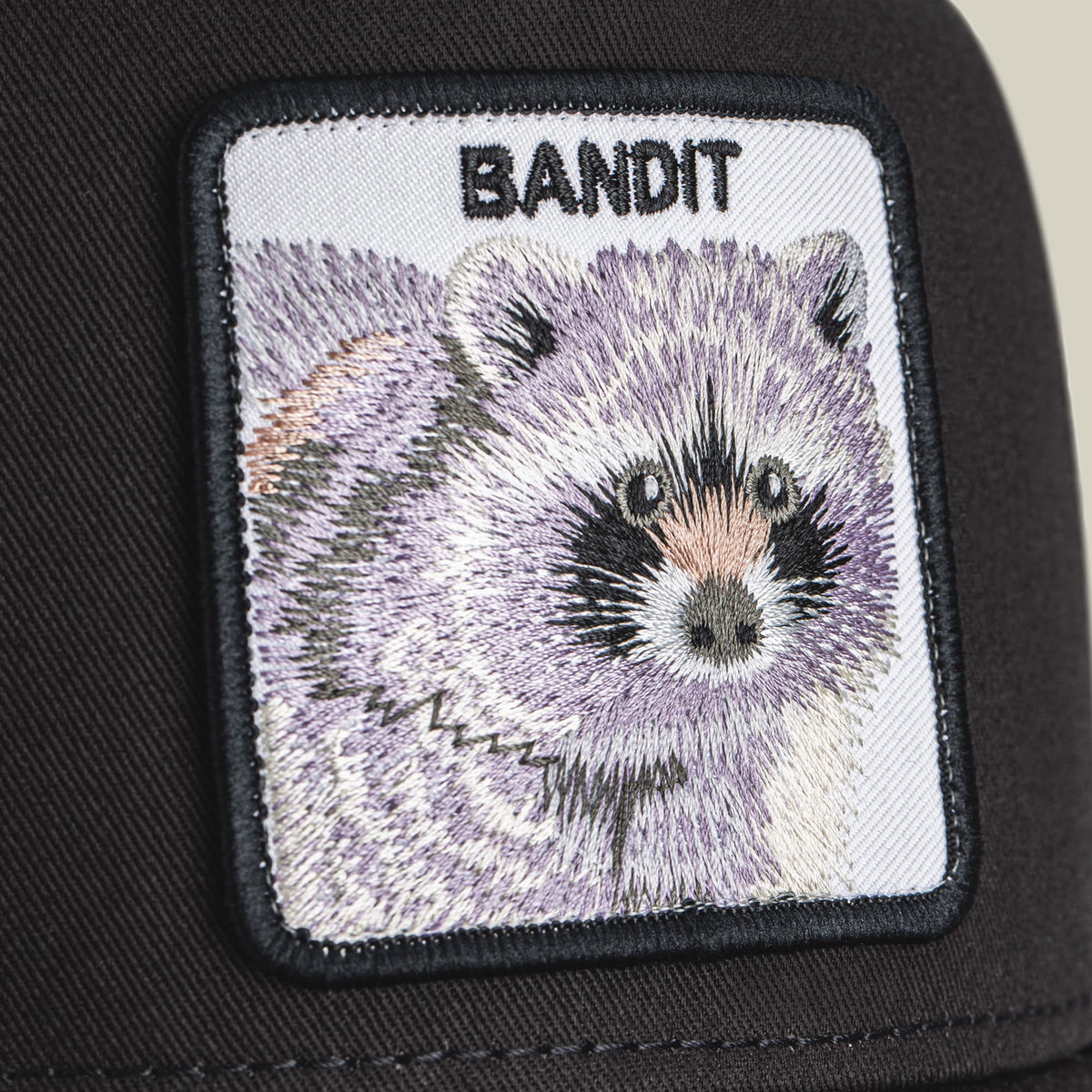 The Goorin Bros. Bandit Trucker Mini is a black trucker hat with an embroidered white patch featuring a detailed raccoon face and the word "Bandit" in black text.