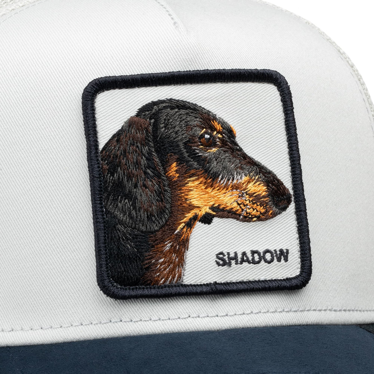 The Suede Shadow Mini trucker hat features a white crown and navy bill with an embroidered dachshund portrait patch labeled "SHADOW" on the front.