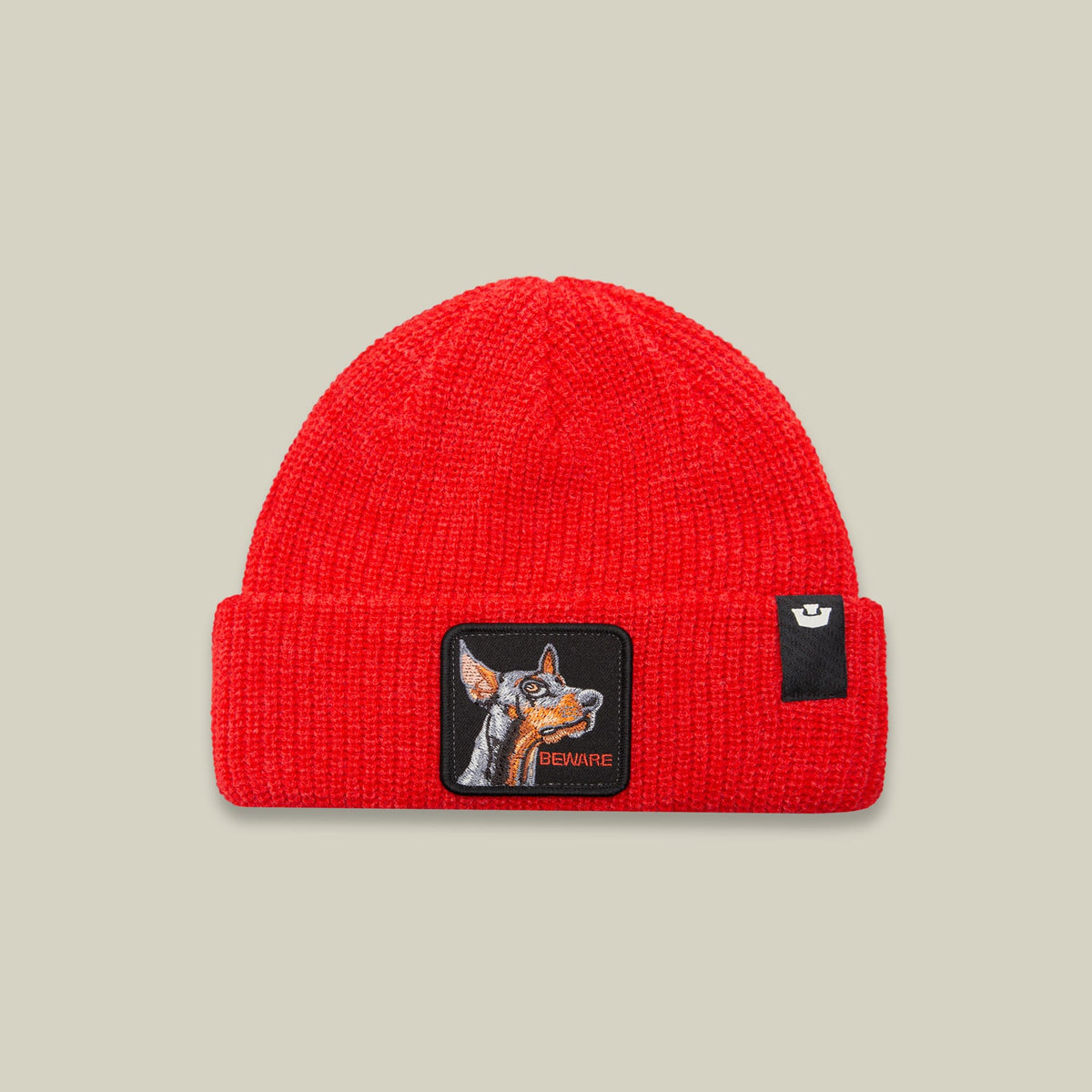 The Beware Beanie Mini by Goorin Bros is a bright red ribbed knit beanie with an embroidered dog patch on the front and a small crown logo patch on the side, shown on a neutral background.