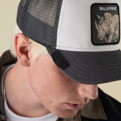 A person with light skin and blonde hair wears a black and white mesh cap featuring a rhino patch and the word BULLETPROOF embroidered on it, paired with the Goorin Bros. Castle CapLoc—a sleek zinc alloy Brim Clip—as a stylish hat accessory.