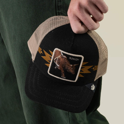 A person holds the Goorin Bros. Canyon Rowdy trucker hat—a black and tan mesh-back cap featuring a front patch with a bull and the word ROWDY—radiating true bull energy. The person is wearing green pants.
