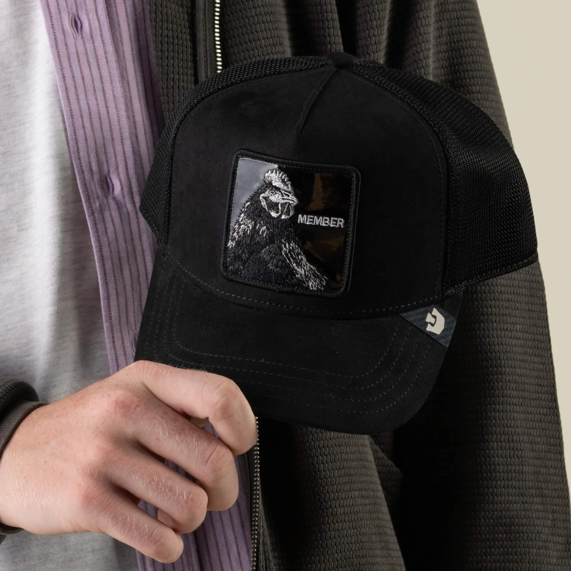 A person holds a jet-black polyester mesh baseball cap, the Black Out Rooster by Goorin Bros., featuring a MEMBER patch with a gorilla illustration on the front.