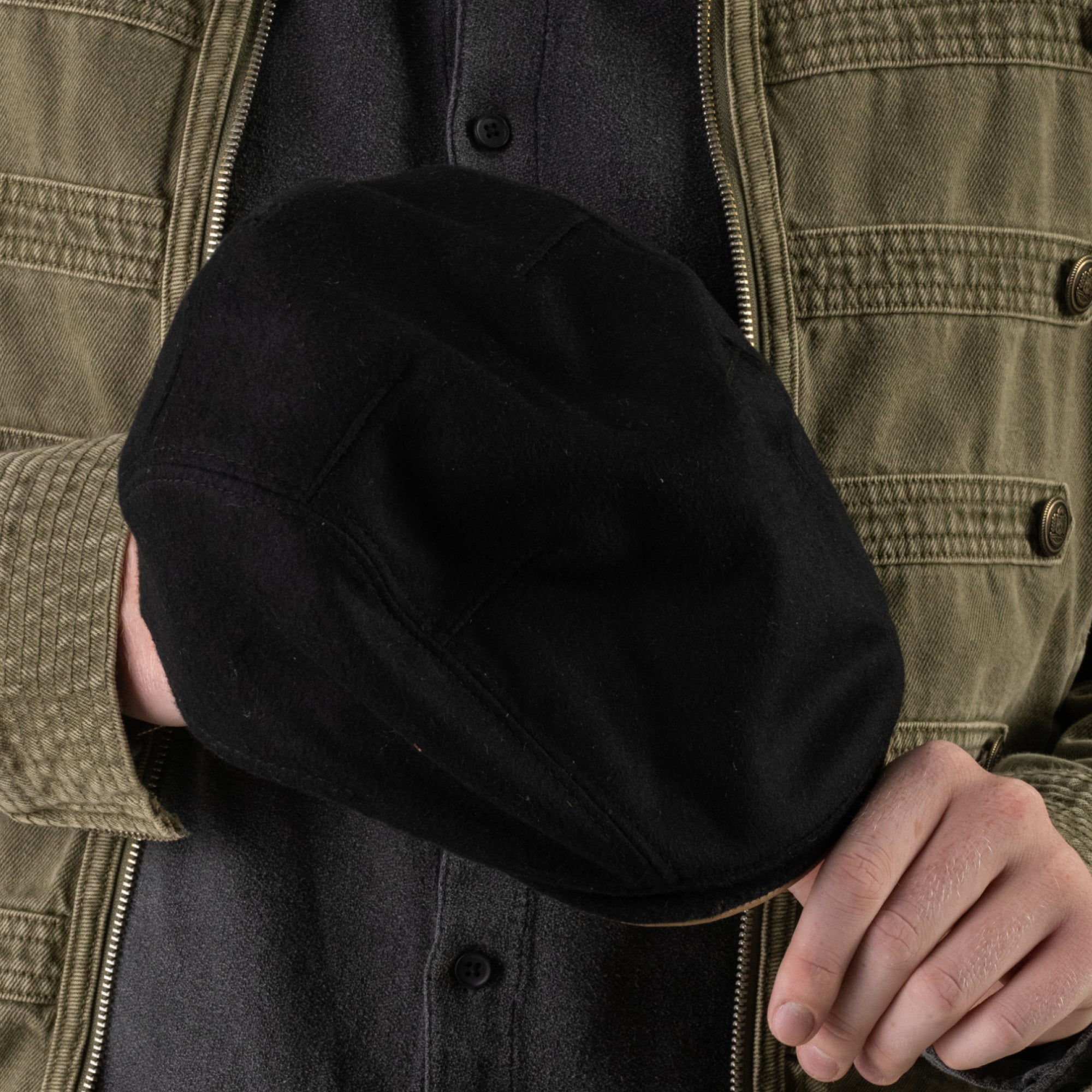 A person in an olive jacket and black shirt holds the Cornerstone black flat cap by Goorin Bros., handmade in New Jersey, in their right hand, with the cap covering part of their chest.