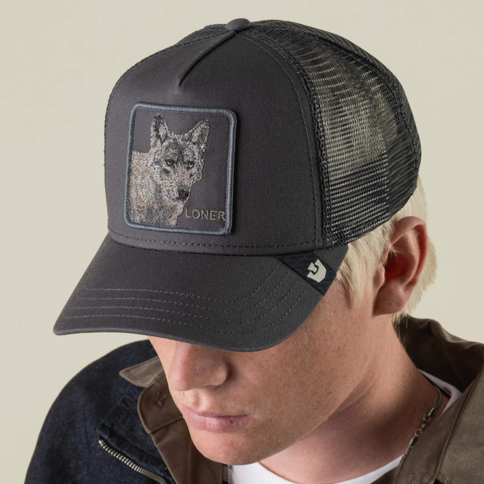 A person wearing the Goorin Bros. “Circle of Life - Loner” black mesh trucker cap, featuring an embroidered wolf patch labeled LONER on the front, is looking down and dressed in a dark jacket.