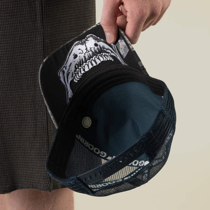 A person holds the Goorin Bros. Bone Head Danger black mesh-back trucker hat, featuring a striking white animal skull graphic printed on the underside of the brim.