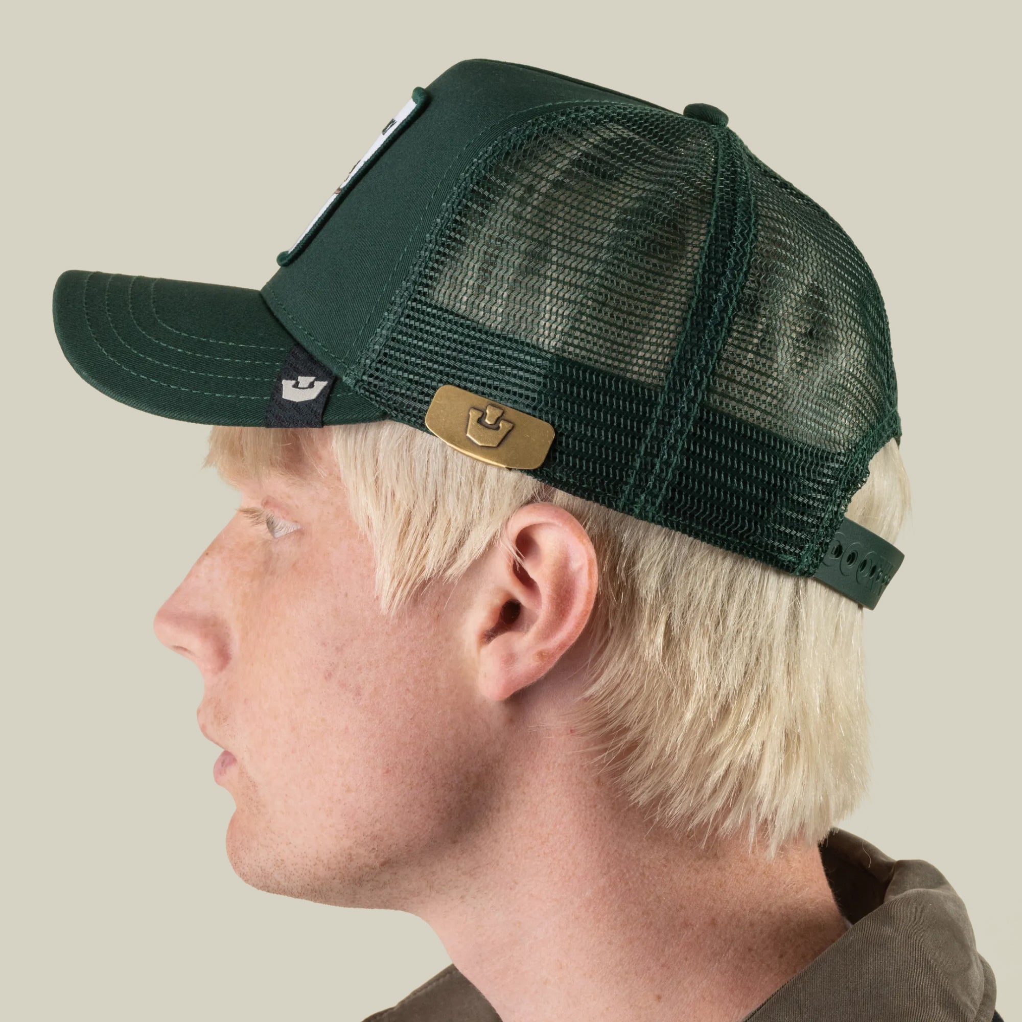 A person with light blond hair is wearing a dark green mesh Goorin Bros. baseball cap, styled with the sleek zinc alloy Castle CapLoc brim clip accessory, seen from the side against a plain background.