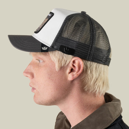 A person with light blond hair is shown in left profile against a plain light background, wearing a Goorin Bros. Castle CapLoc black and white mesh trucker hat featuring a zinc alloy Brim Clip, along with a brown collared jacket.