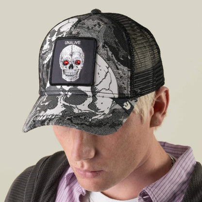 A person with blond hair is looking downward while wearing the Goorin Bros. Bone Head Unalive trucker hat, which features a black and white camo design, a skull patch labeled “UNALIVE” with red eyes, and a breathable mesh back.
