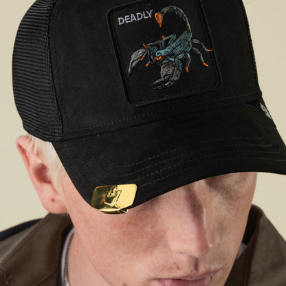 A person is wearing a black Goorin Bros. Castle CapLoc cap featuring a scorpion patch labeled DEADLY, with a gold zinc alloy Brim Clip accessory attached to the brim. The person is looking down, and their face is partially visible.